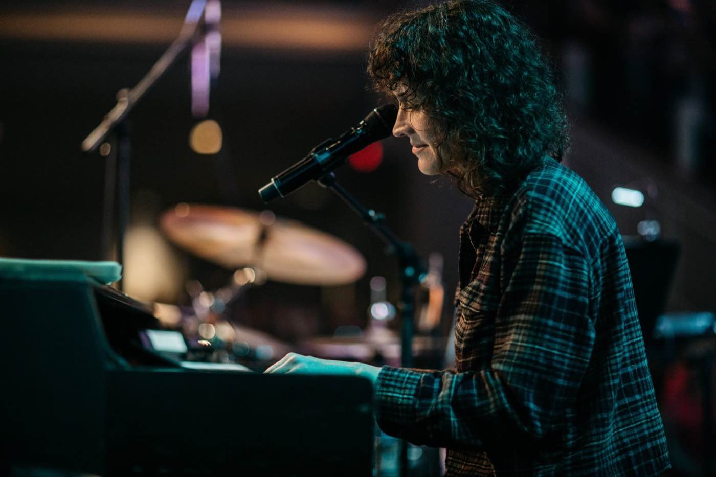 Side profile of a musician with curly hair playing a keyboard on a dimly lit stage.