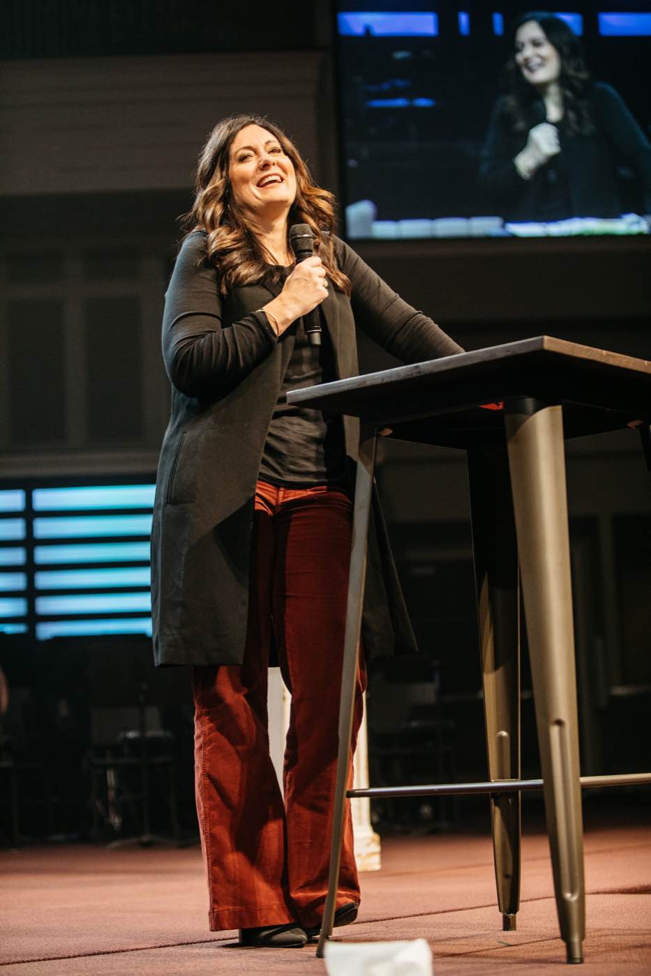 Woman speaking at a podium with a microphone, smiling brightly under stage lights.