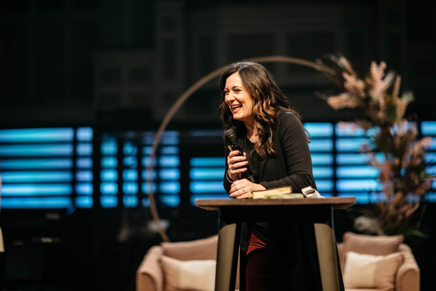 Woman speaking at a podium, laughing, with blue stage lights and warm decor in background.