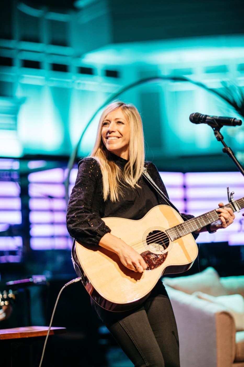 Smiling female musician playing acoustic guitar on stage with vibrant blue and purple lighting.