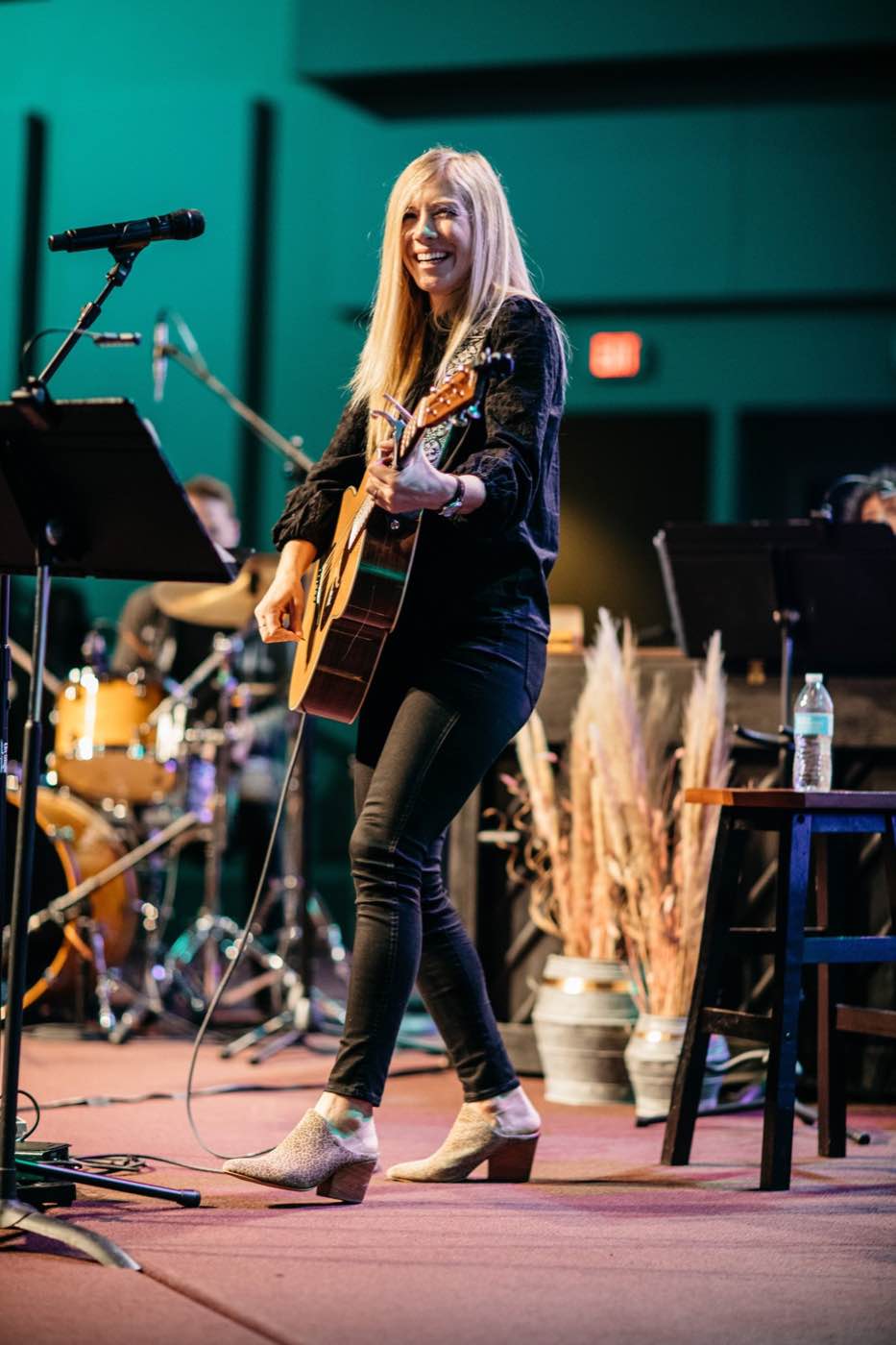 Energetic female musician smiling and playing acoustic guitar on stage under bright teal lights.