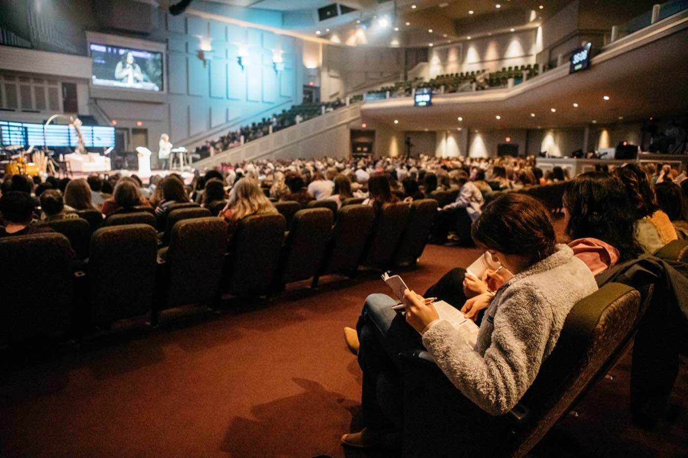 Wide shot of a large audience in a dimly lit auditorium, focused on a stage with a screen.
