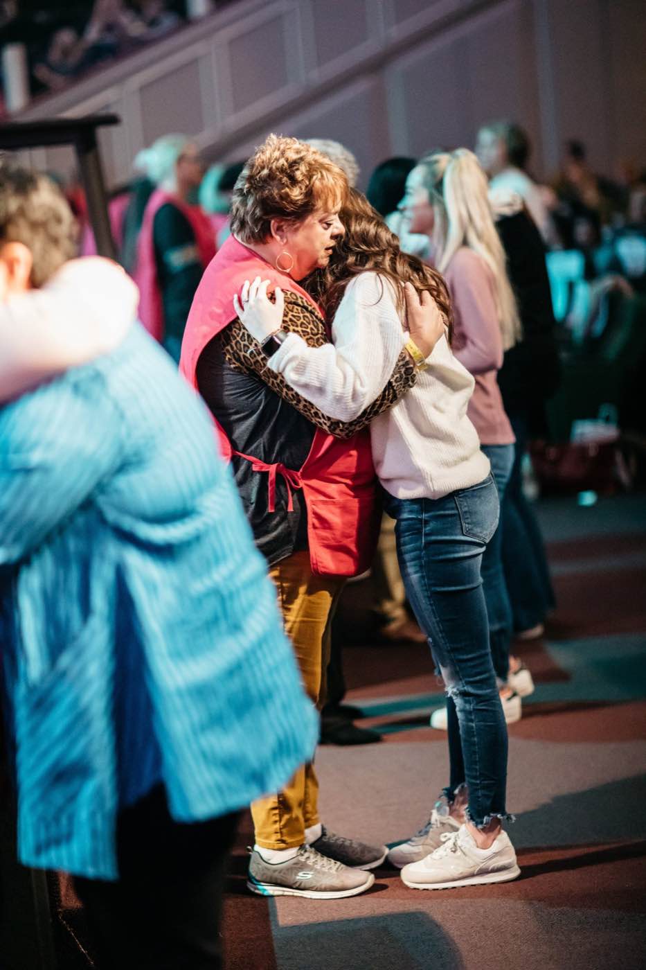 Two women embrace warmly in a dimly lit indoor setting, surrounded by a blurred crowd.