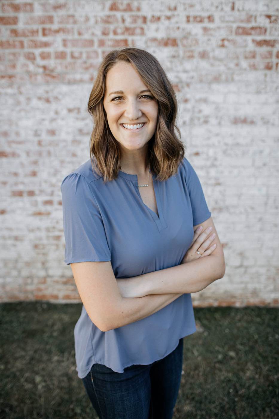 Smiling woman in blue top with arms crossed against a brick wall, natural light
