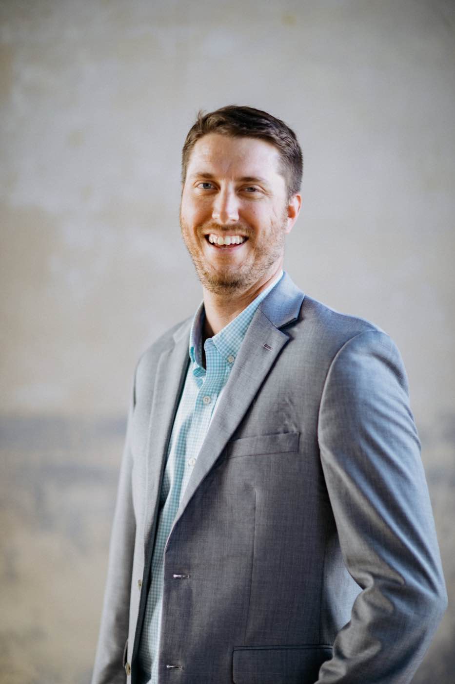 Smiling man in a grey suit and blue checkered shirt against a light grey wall, well-lit.