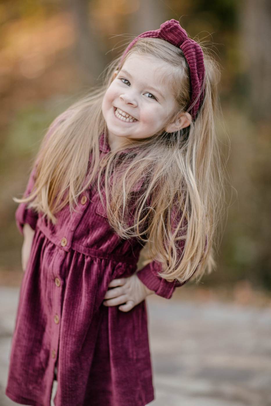 Close-up portrait of a joyful young girl with long blonde hair and a maroon dress, smiling brightly outdoors in soft, natural light.