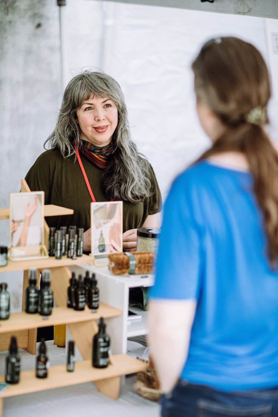 A smiling vendor with long gray hair and a colorful scarf stands behind a display of natural products, engaging with a customer.