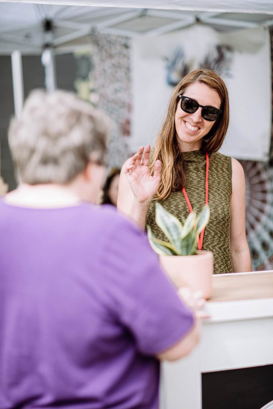 Smiling woman in sunglasses waves at an event, warm natural light, joyful mood.
