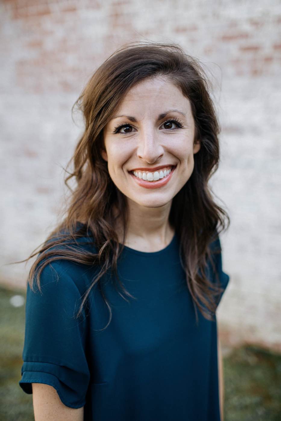 Close-up portrait of a smiling woman with long brown hair against a soft-focus brick wall, natural light.