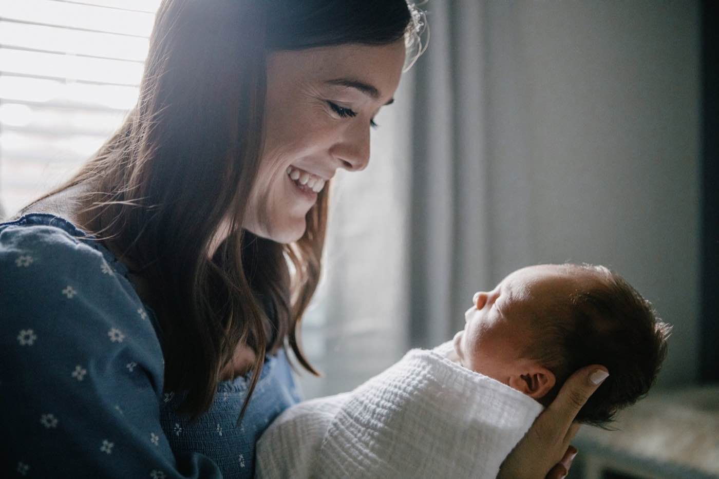 A mother smiles lovingly at her newborn baby, bathed in soft, natural light, creating a tender moment.
