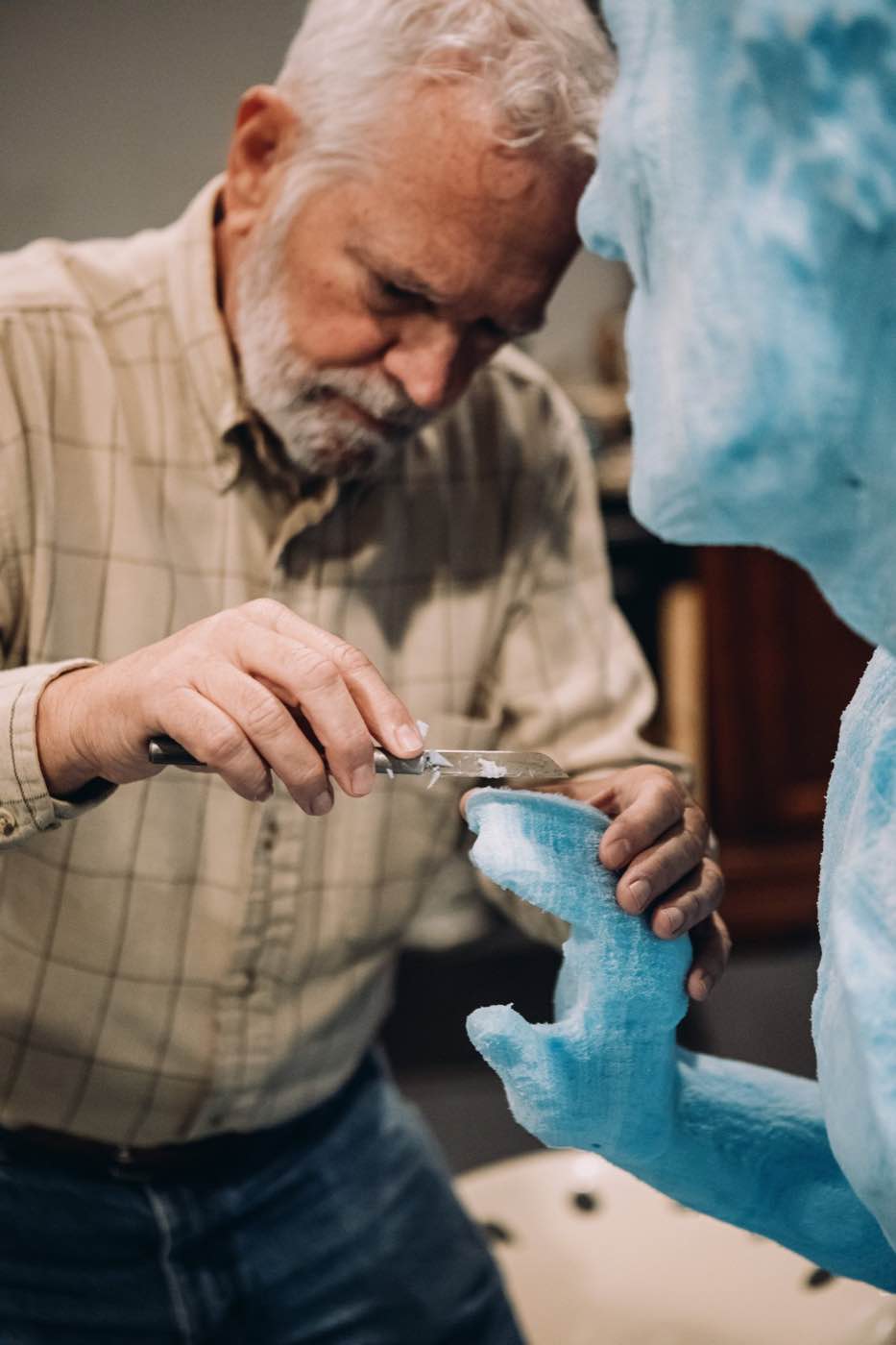Close-up of an older man with a white beard carving a light blue sculpture with a knife.
