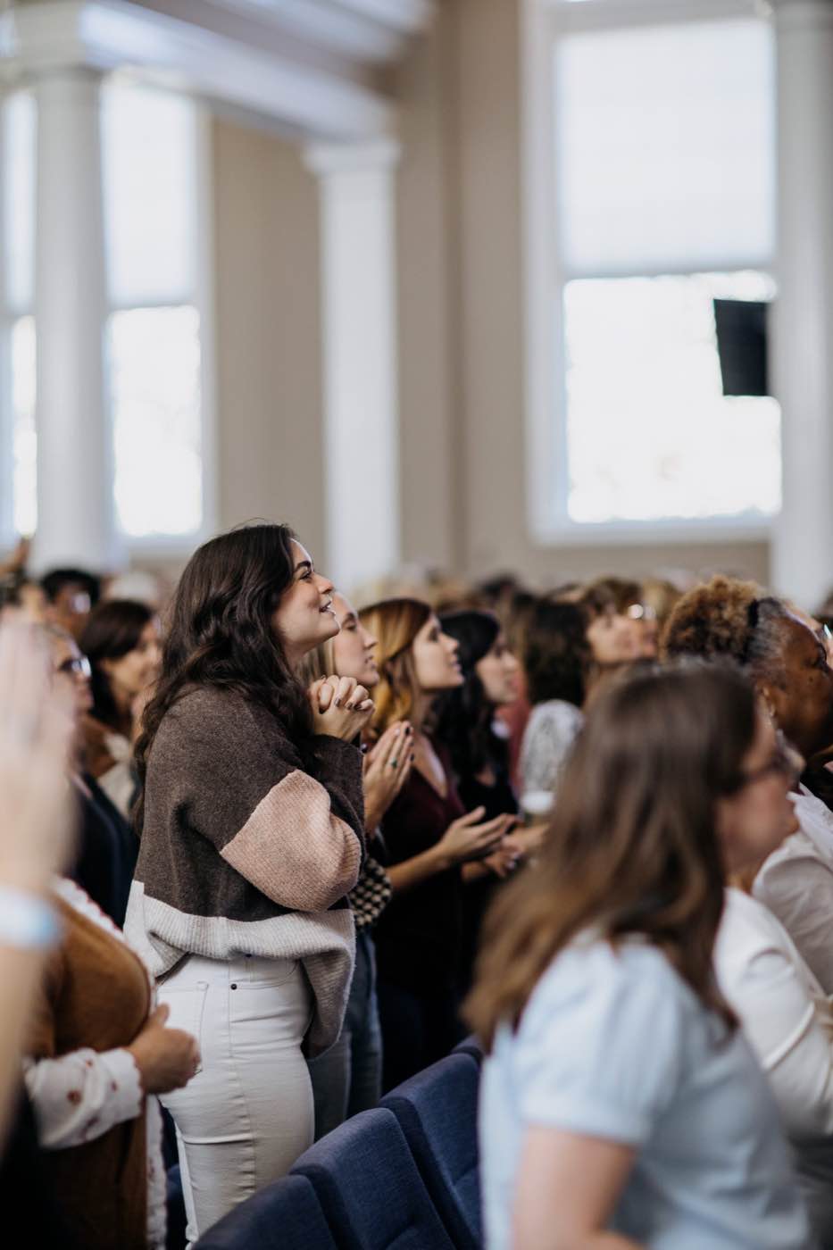 A woman with dark hair, eyes closed and hands clasped, stands in a sunlit room with other people, creating a serene and spiritual mood.