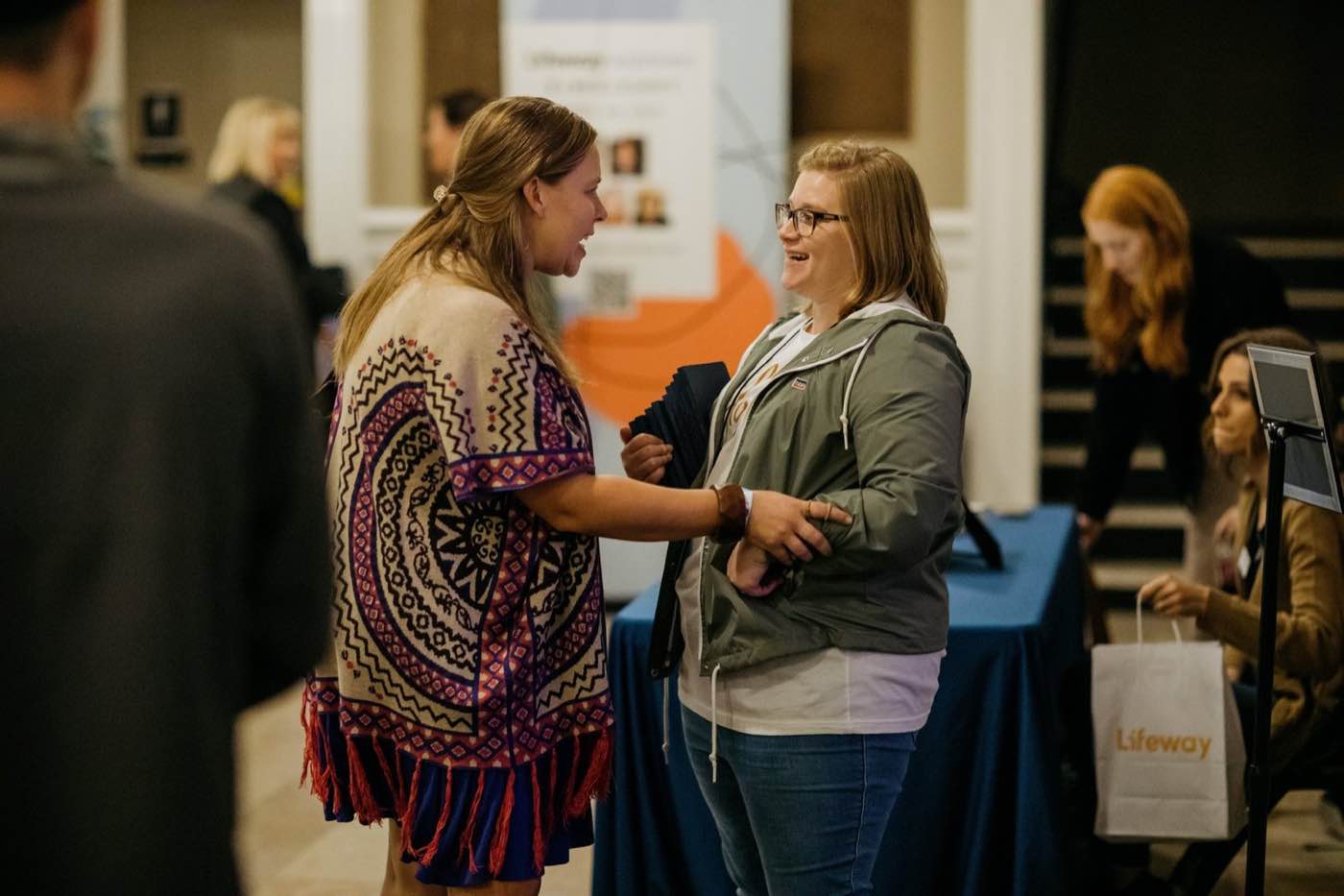 Two women smiling and talking at an indoor event, warm lighting, lively mood.