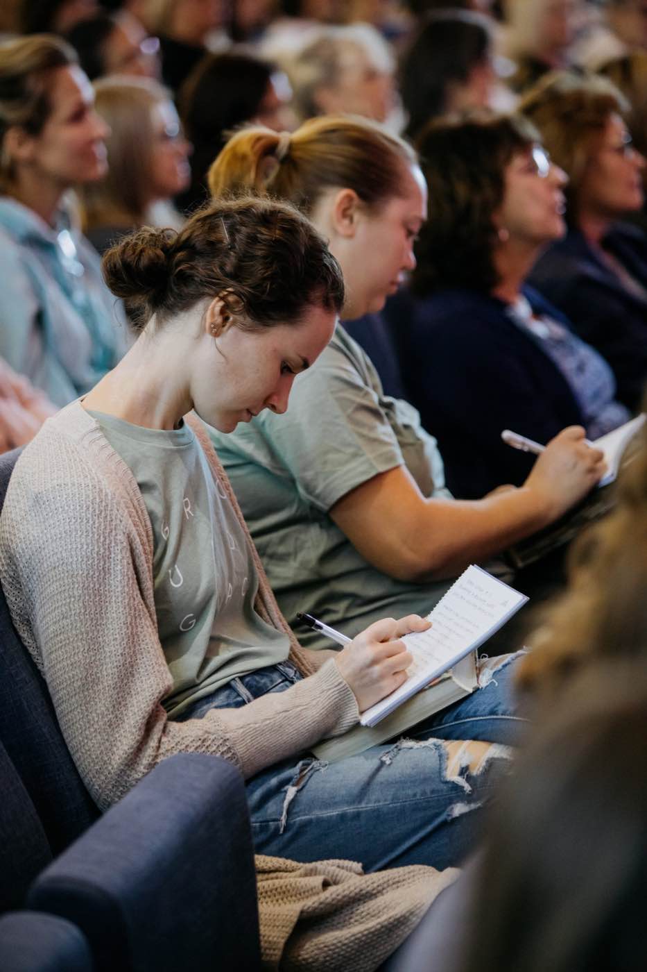 Attendees at a conference or seminar, focused on taking notes in a warm, natural light setting.