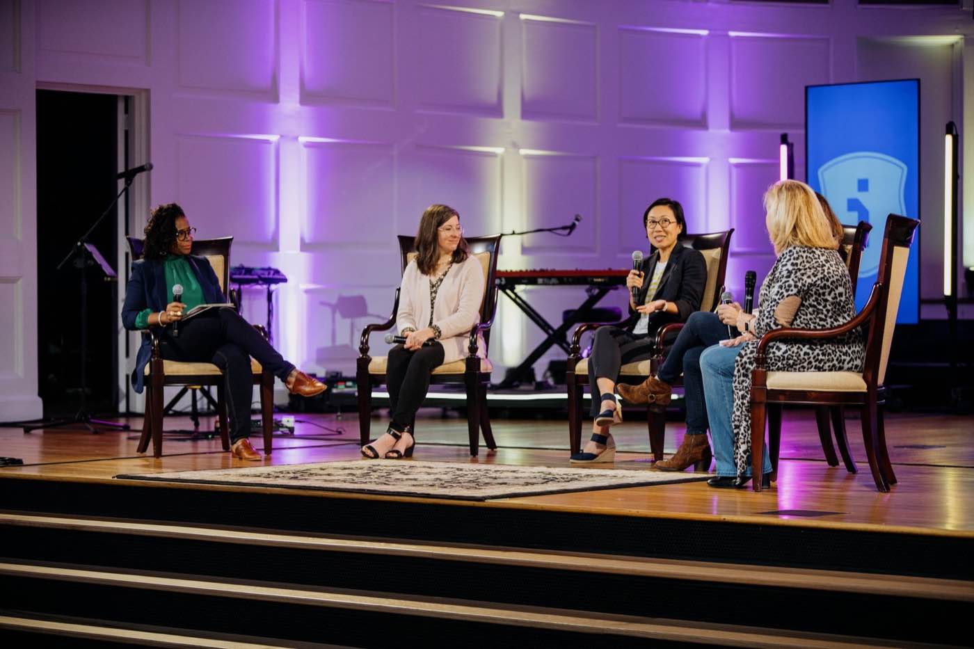 Four women on stage, seated in chairs, engaged in a panel discussion under purple lighting.
