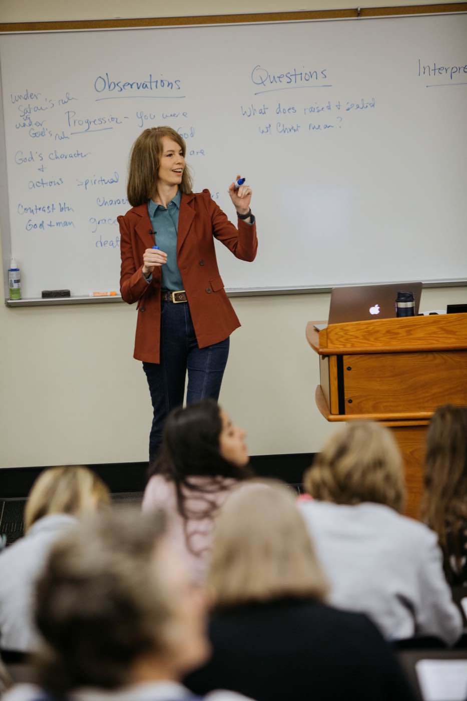 A female speaker in a classroom, smiling and gesturing while holding a marker, with a whiteboard behind her.