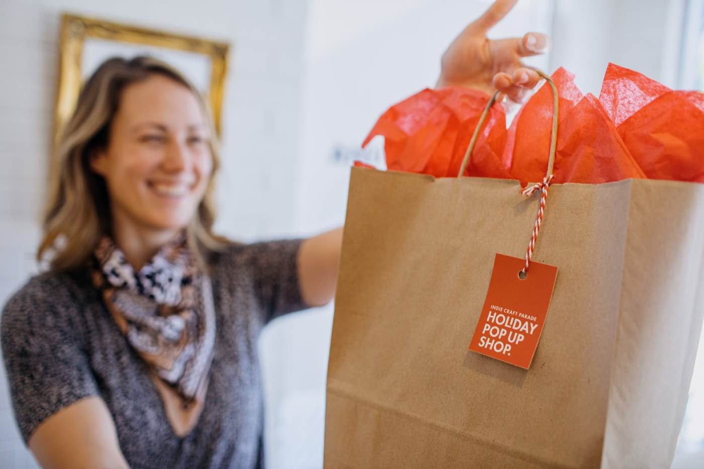 Smiling woman holding a brown paper bag with red tissue paper and a 'Holiday Pop Up Shop' tag.