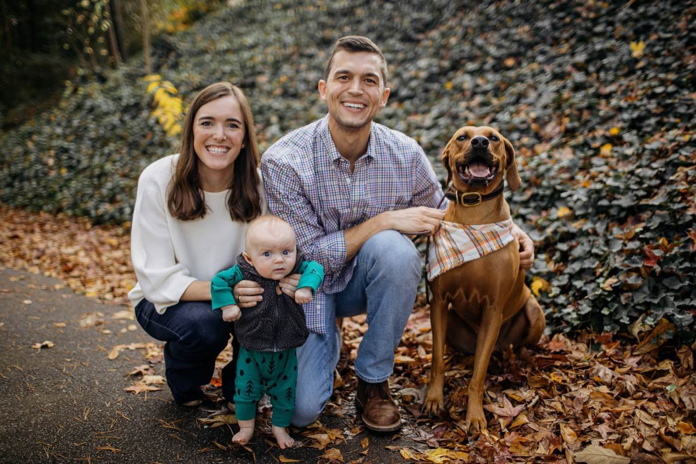 A joyful family of three and their dog pose outdoors on a path with autumn leaves, natural light.