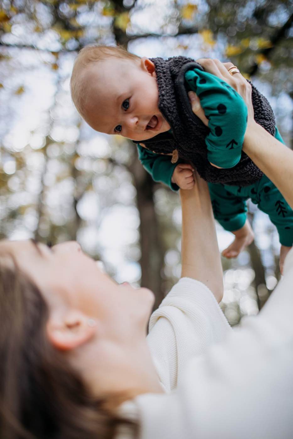 Joyful mother holding baby up in the air against a bokeh background of trees, natural light