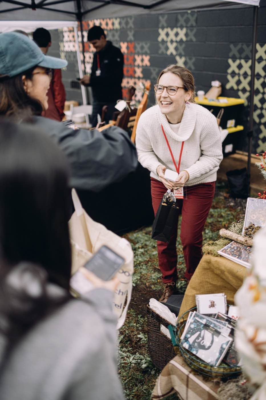 Joyful woman in glasses and a white sweater laughing at an outdoor market stall.