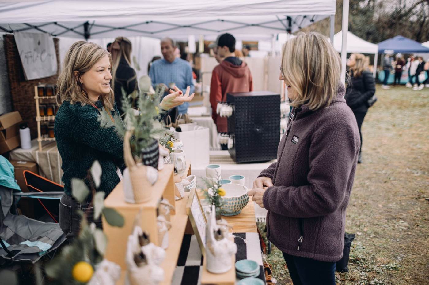 Two women interacting at an outdoor market stall under a white tent, natural light.