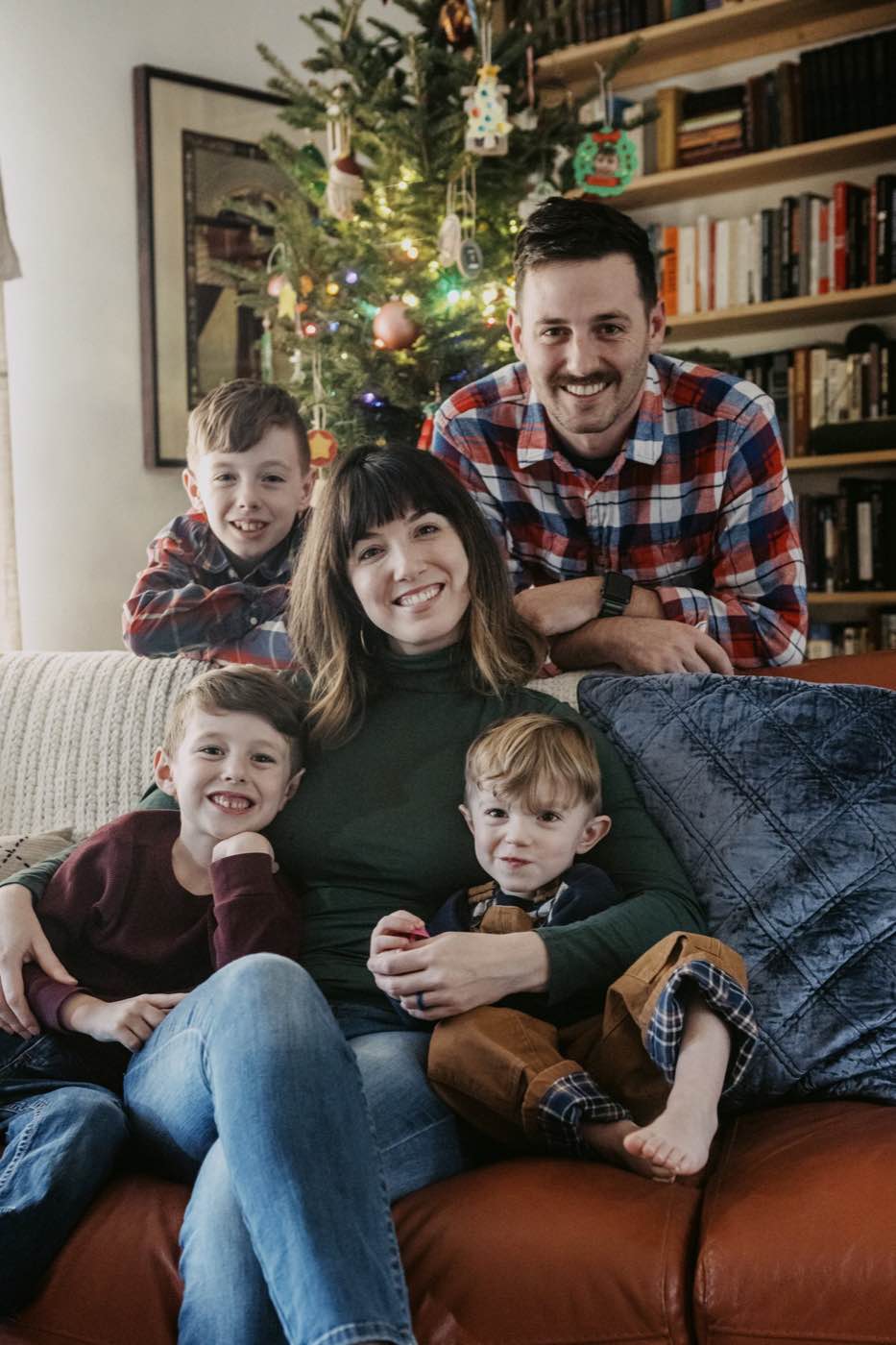 A joyful family of five, including three young boys, smiles at the camera in a warmly lit living room with a Christmas tree.