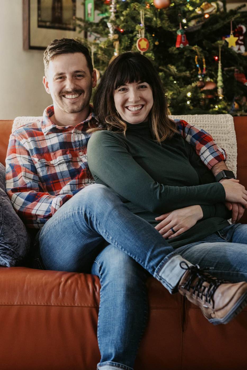 Joyful couple smiling on a brown leather couch with a decorated Christmas tree in the background, warm indoor lighting.