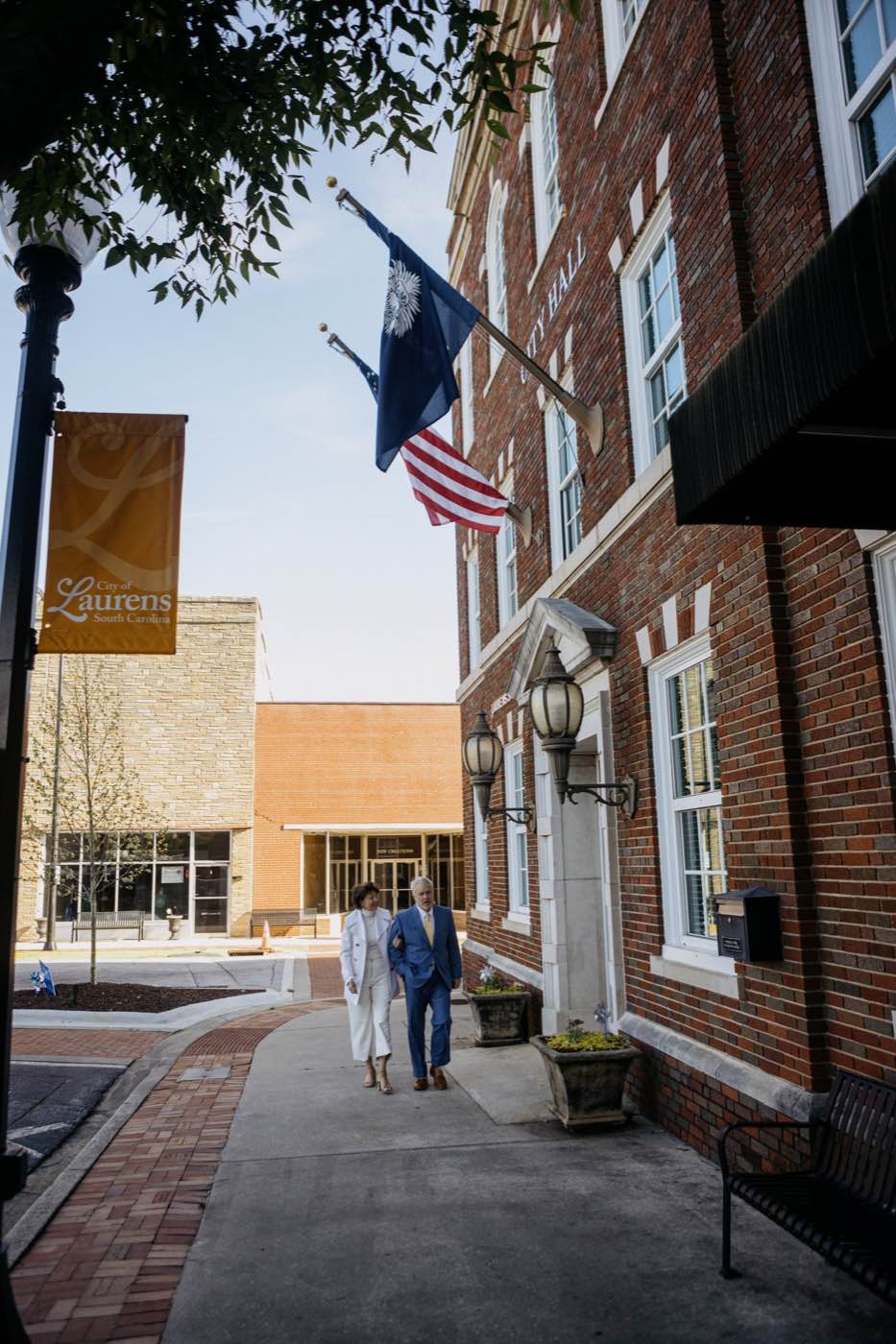 Bright outdoor shot of an older couple walking arm-in-arm past a brick city hall building.