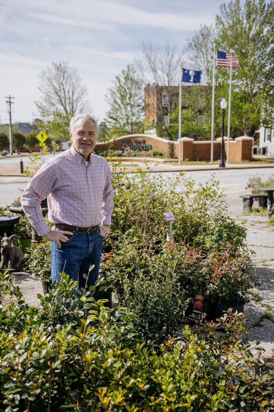 Man smiling in a garden center with a town sign and flags in the background under bright daylight.