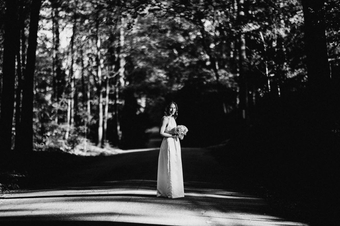 Dramatic black and white portrait of a woman in a long dress holding flowers on a shaded road.