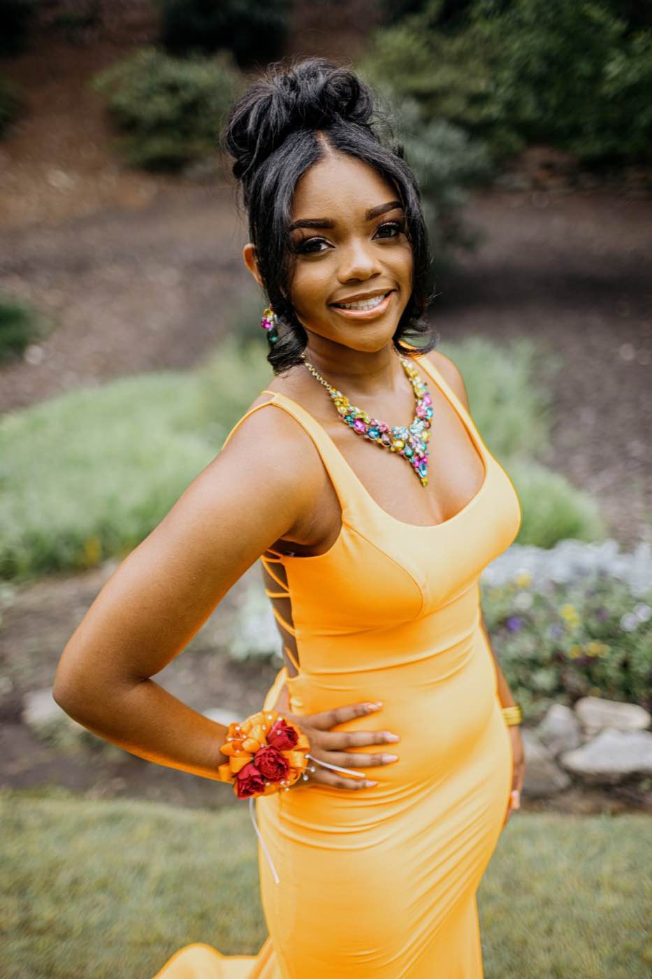 Smiling woman in a vibrant yellow dress and colorful jewelry, standing outdoors in soft natural light.