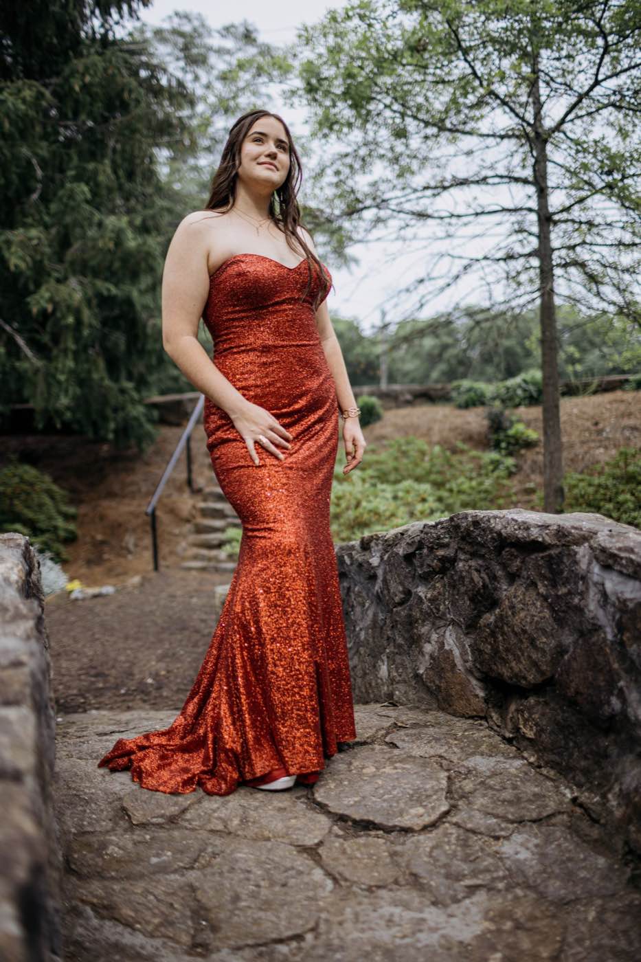 Young woman in a shimmering red gown stands on a stone path, looking up with a serene expression amidst lush greenery.