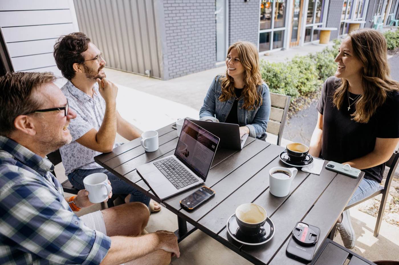 Four colleagues smiling and chatting at an outdoor cafe table with laptops and coffee, bright daylight.