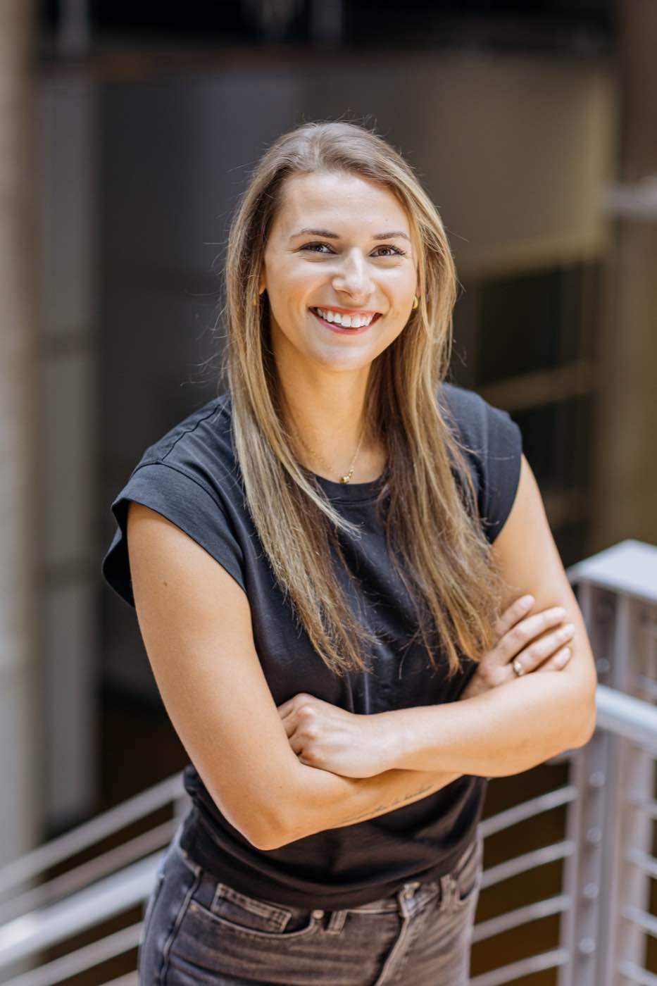 Smiling woman with long blonde hair, arms crossed, in a modern indoor setting with soft lighting.