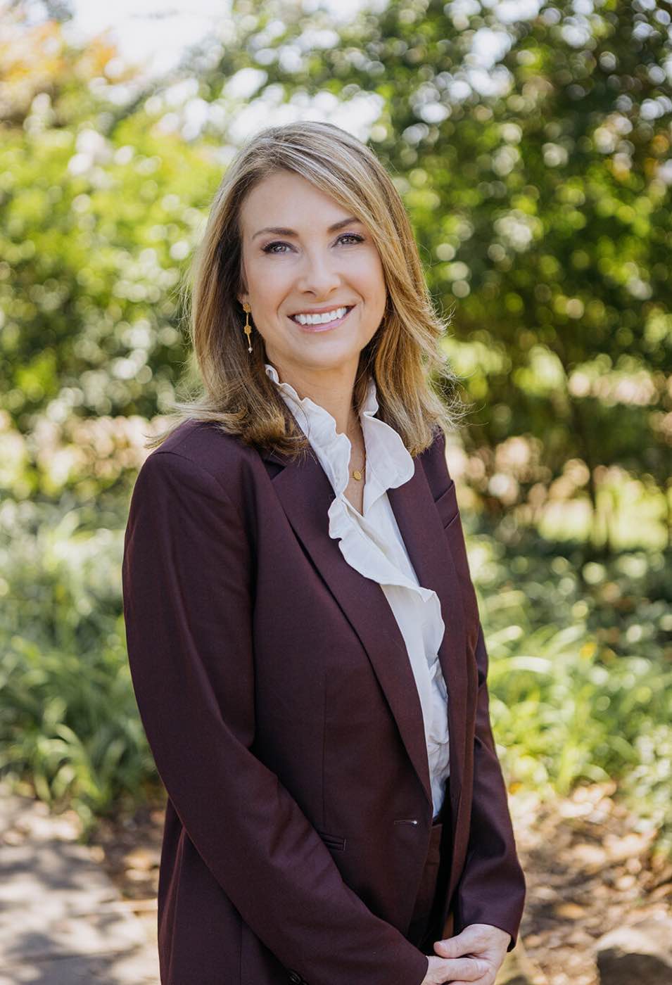 Professional woman in a maroon suit and white ruffled blouse smiling outdoors with soft natural light.