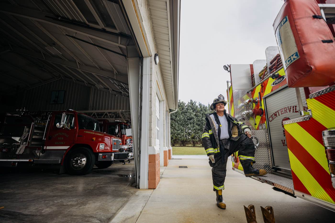 Firefighter in full gear, one foot on a fire truck, looking at the camera with a smile.