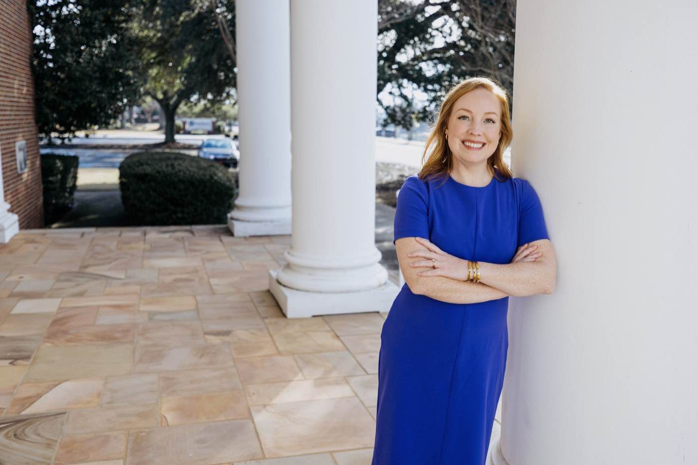 Smiling woman in blue dress leaning against a white pillar on a sunny day.
