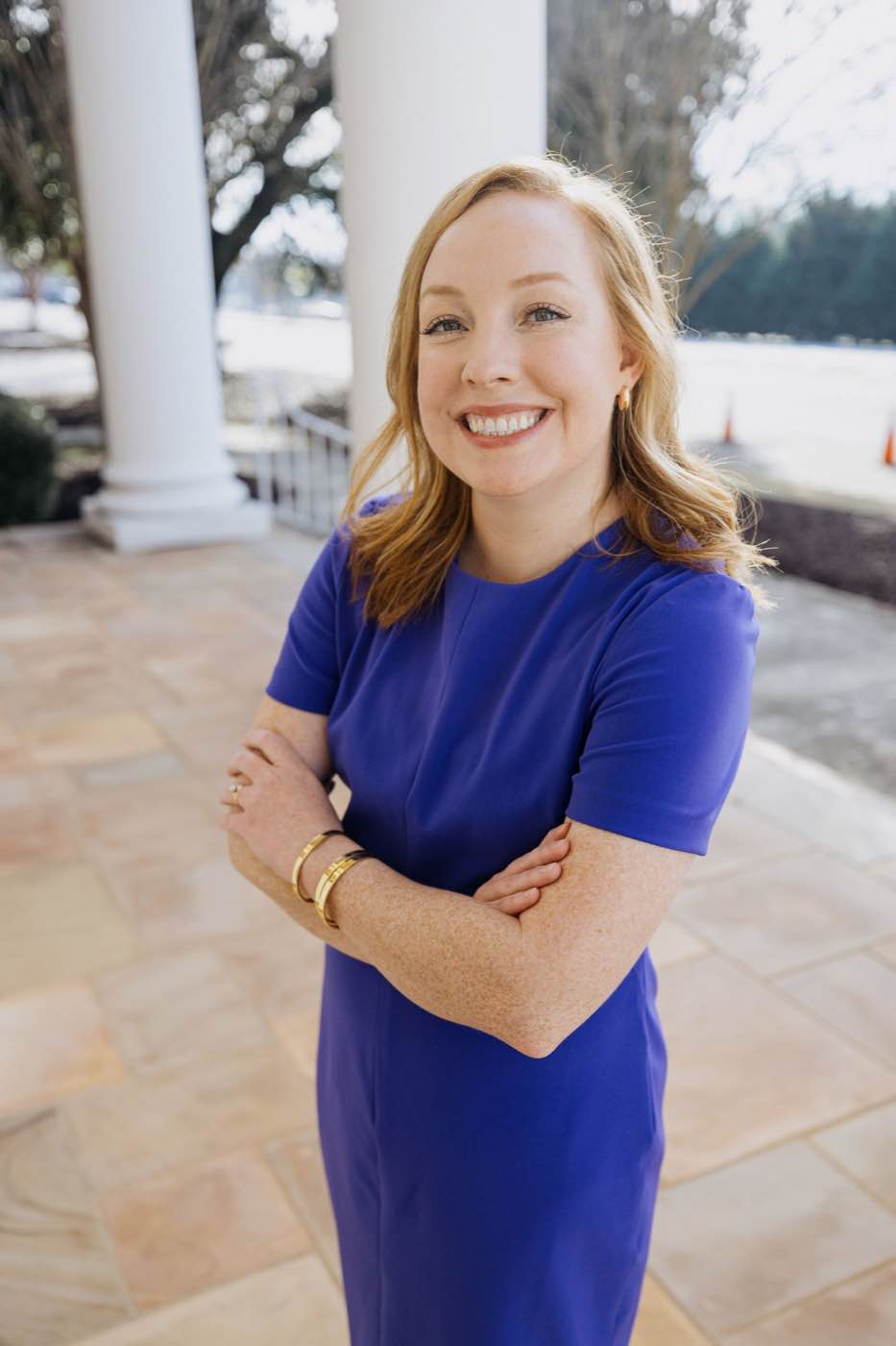 Smiling woman in blue dress with arms crossed, standing outdoors in natural light.