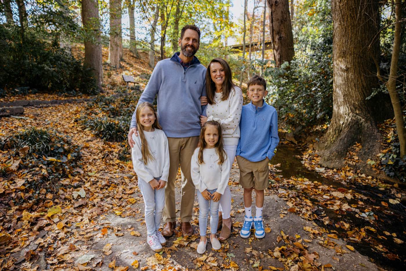 A joyful family of five stands on a path covered in autumn leaves, bathed in warm, natural light.