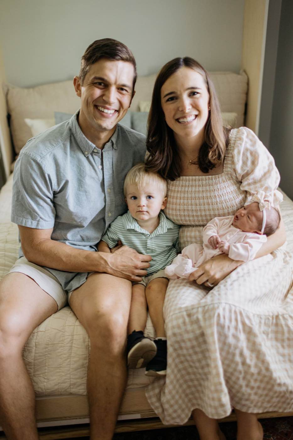 A joyful family of four, including a newborn, sitting on a bed in soft, natural light.