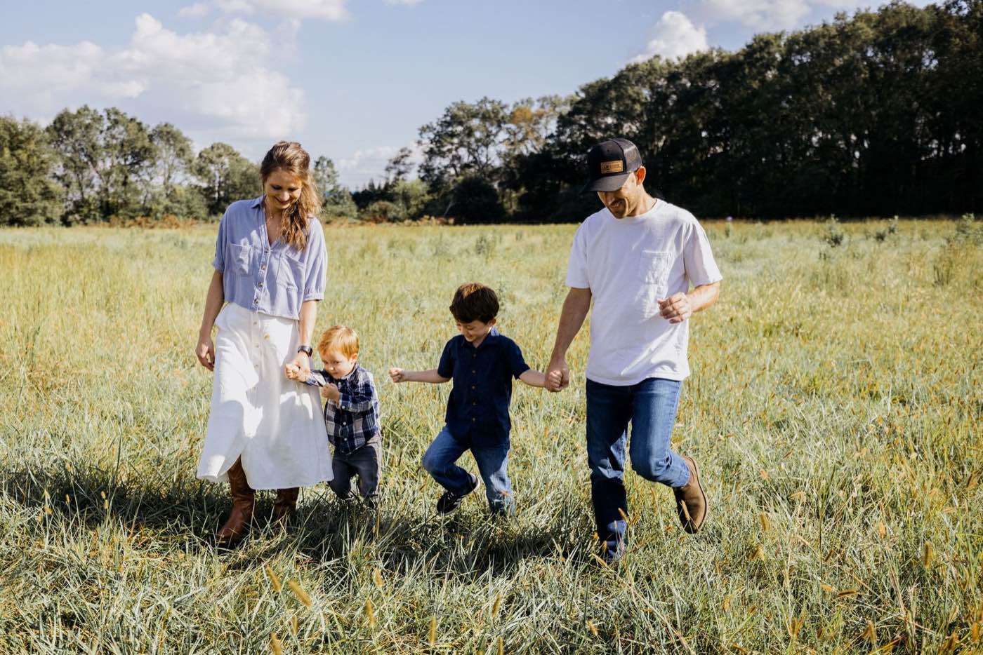 A family of four, parents and two young boys, walk hand-in-hand through a sunlit grassy field on a clear day.