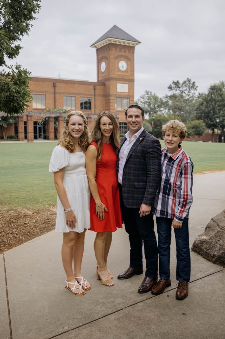 A family of four smiling outdoors in front of a brick building with a clock tower on a cloudy day.