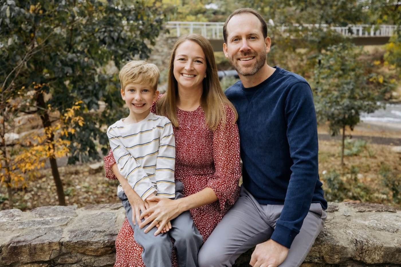 A joyful family of three, a man, woman, and young boy, smiling brightly in an outdoor setting with natural light.