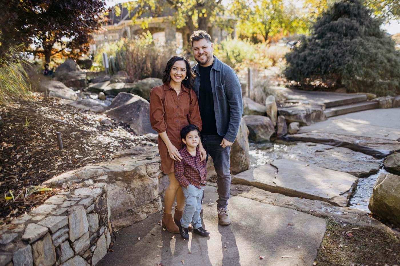 A family of three smiling at the camera in a park with natural light and warm tones.