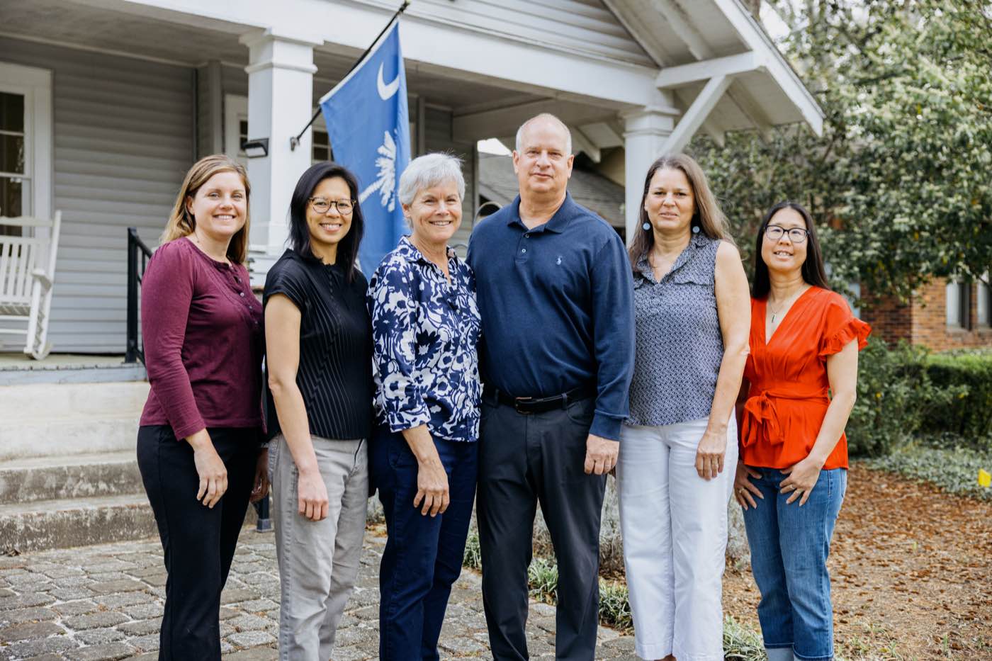 Six smiling colleagues stand outdoors in front of a building with a flag, natural light.